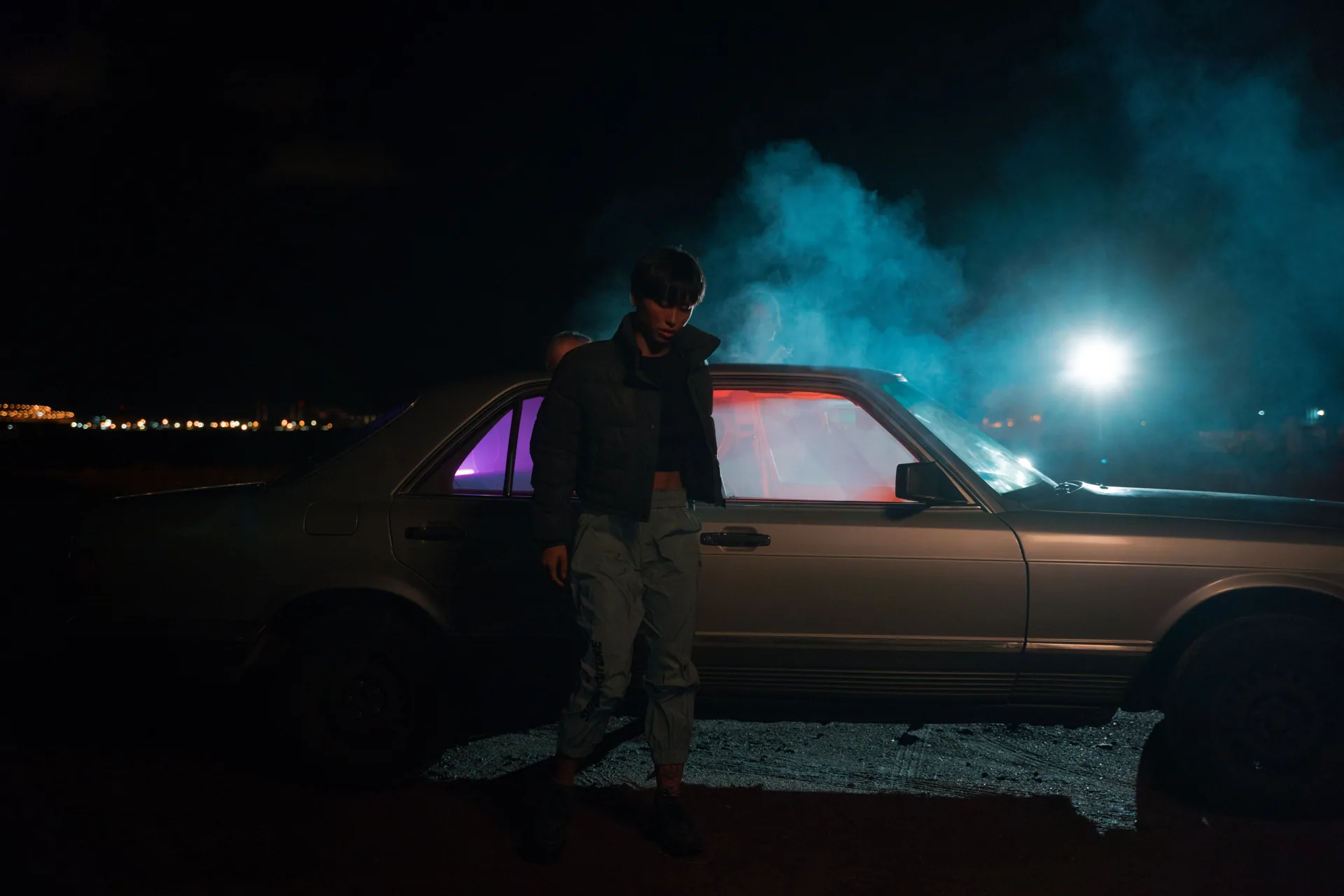 A figure beside a car under blue night lighting.